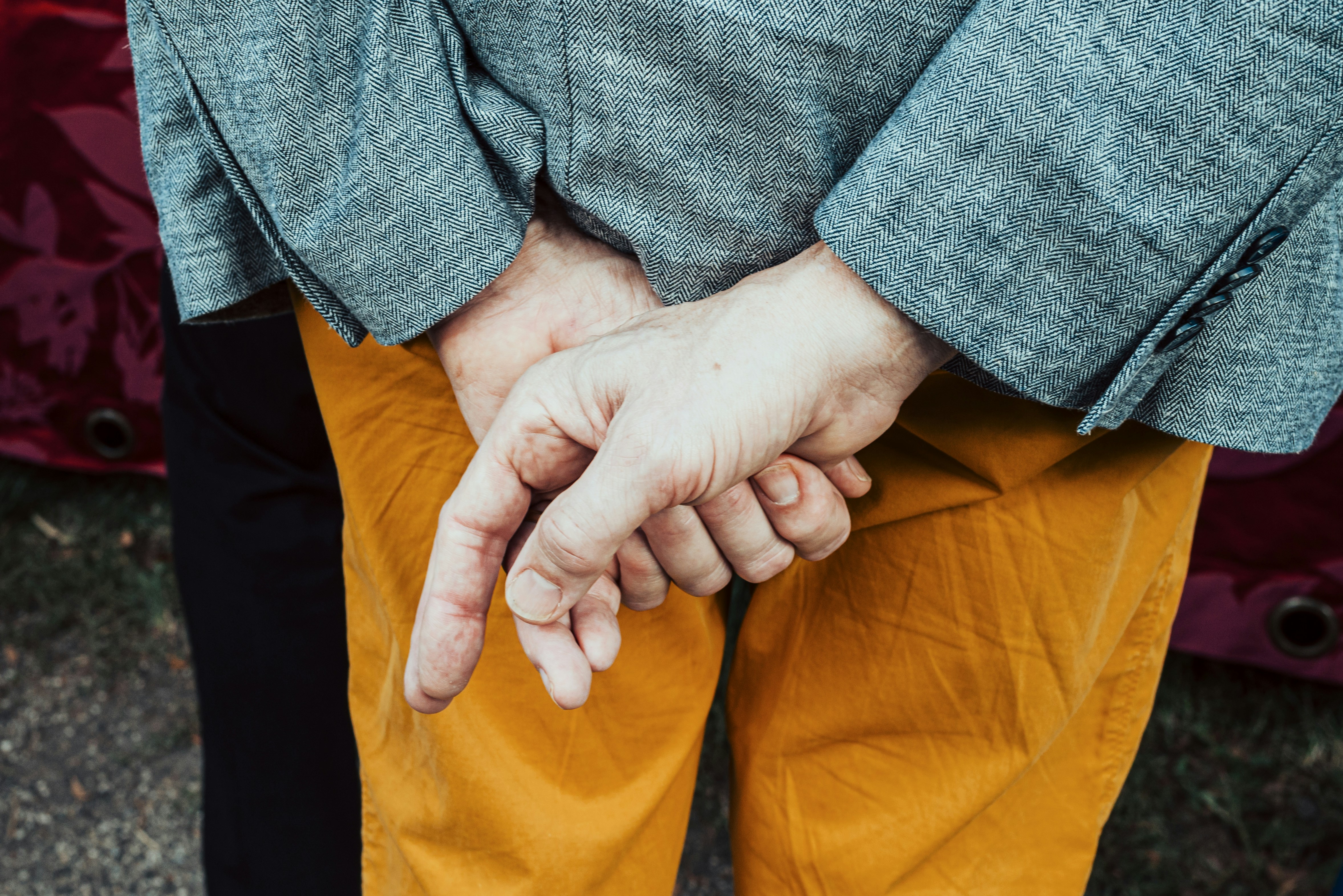 A person with hands clasped in front, wearing a grey herringbone blazer and mustard trousers